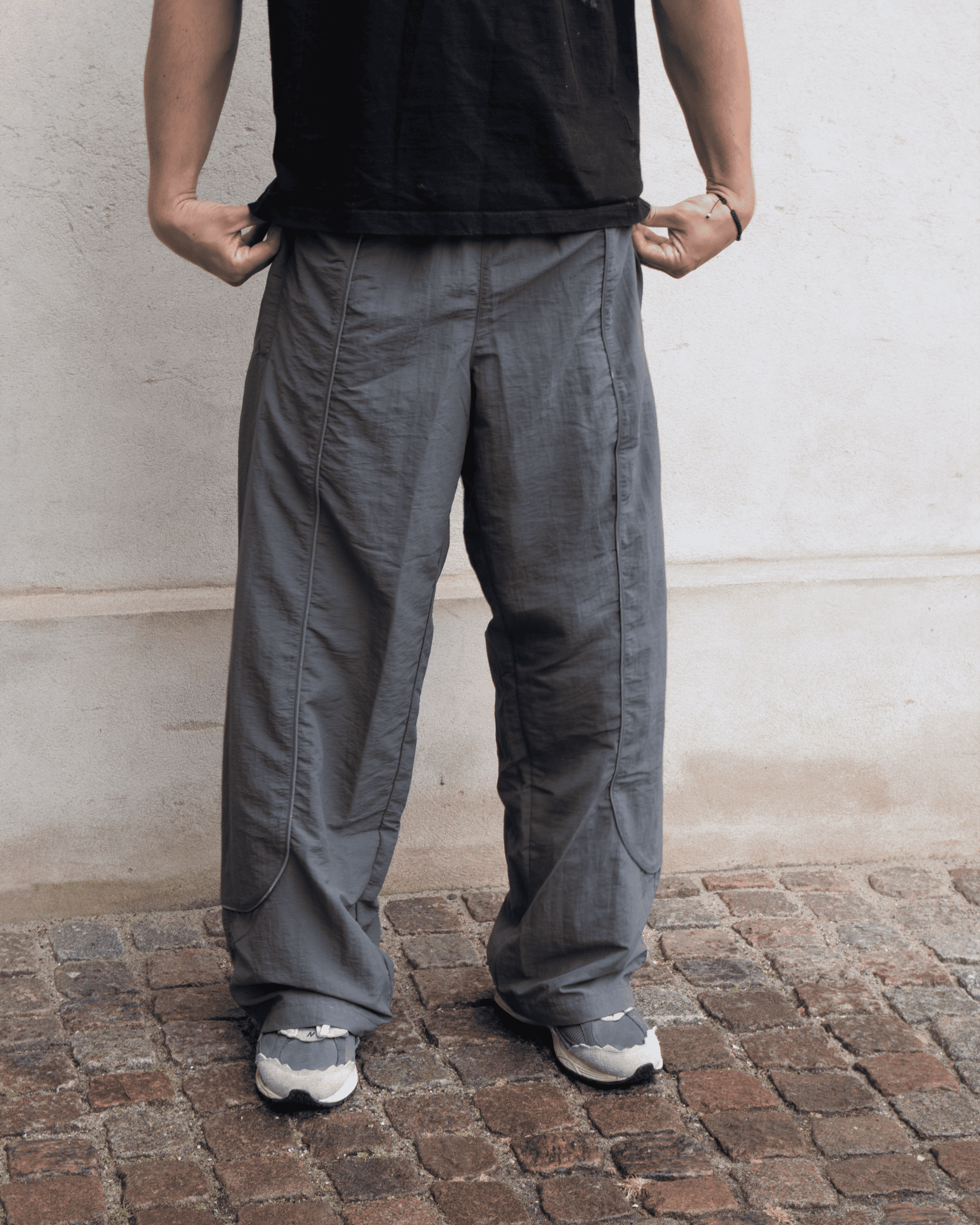 Lifestyle shot of our Track Pants Steel, in a daylight setting up against an ecru colored  wall and brick flooring